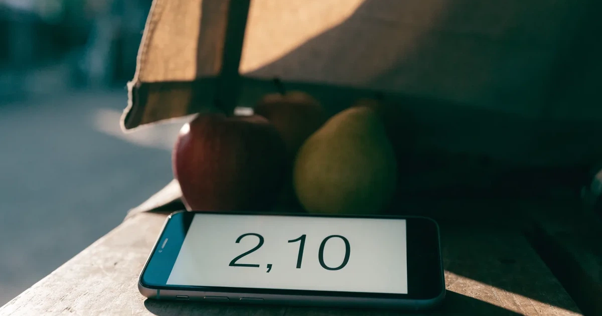 Ein Smartphone mit dunklem Display liegt auf dem Holztisch eines Marktstands zwischen Äpfeln und Birnen, morgendliches Licht fällt durch eine Plane.