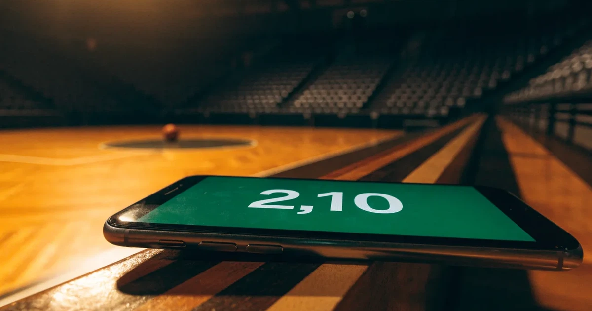 Ein Basketball liegt auf dem hellen Parkett einer leeren Halle, im Hintergrund steigen die Tribünenreihen im Halbdunkel an.
