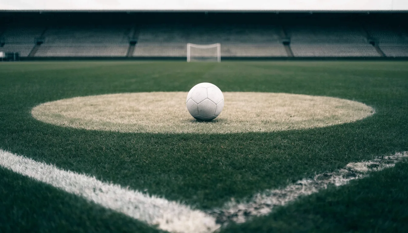 Ein Fussball liegt auf dem Anstosspunkt eines nassen Rasens, dahinter eine leere Tribüne unter grauem Himmel.