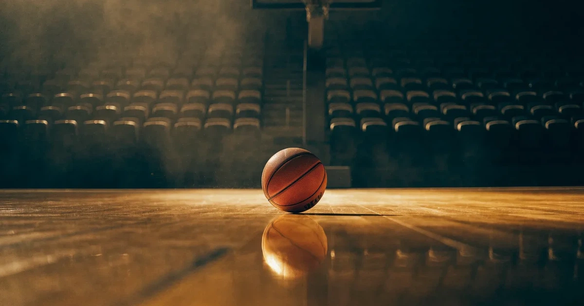 Ein orangefarbener Basketball liegt auf dem Parkett einer leeren Halle unter hellem Flutlicht, die Tribünen sind dunkel.