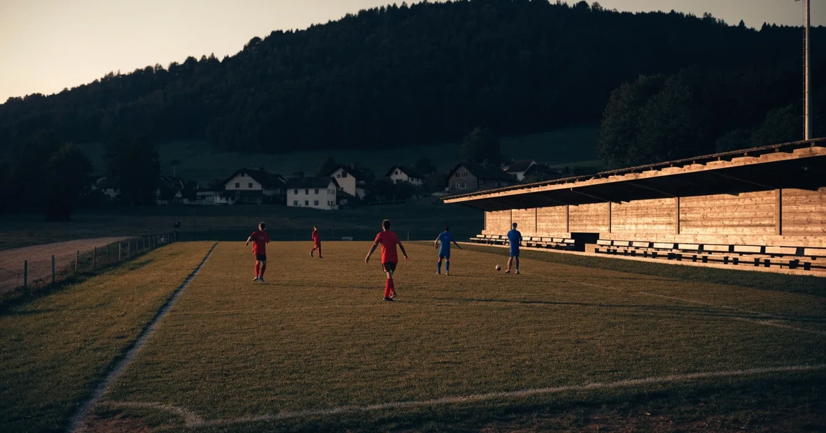Ein kleiner Fussballplatz mit einfacher Holztribüne und Rasenstreifen am Rand einer Ortschaft, im Hintergrund ein bewaldeter Hügel im Abendlicht.