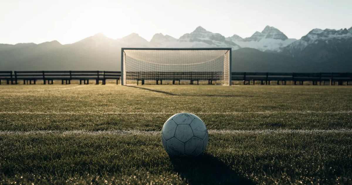 Ein Fussball liegt am frühen Morgen auf dem Rasen eines leeren Fussballfeldes, dahinter eine kleine Tribüne und schneebedeckte Berge.