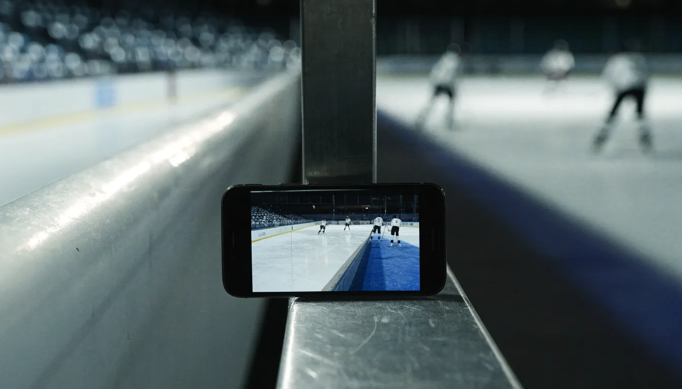 Ein Eishockeyschläger lehnt an der Bande einer leeren Halle, auf dem Eis spiegeln sich die Deckenlampen.